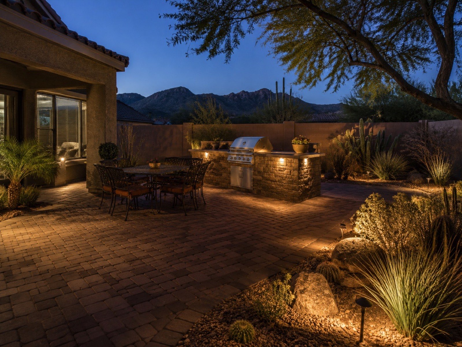 Outdoor kitchen with built-in grill, dining area, and landscape lighting on a paver patio at dusk in Tucson